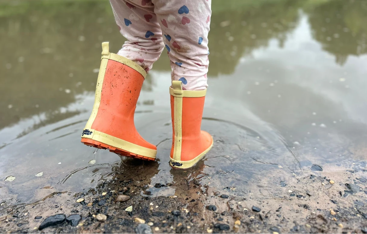 Child wearing orange WelliePaws boots splashing through a puddle